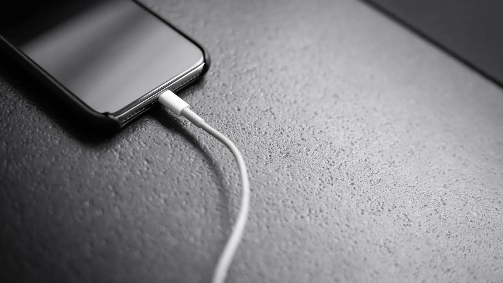 A black smartphone lying on a grey table with a white charging cable plugged in