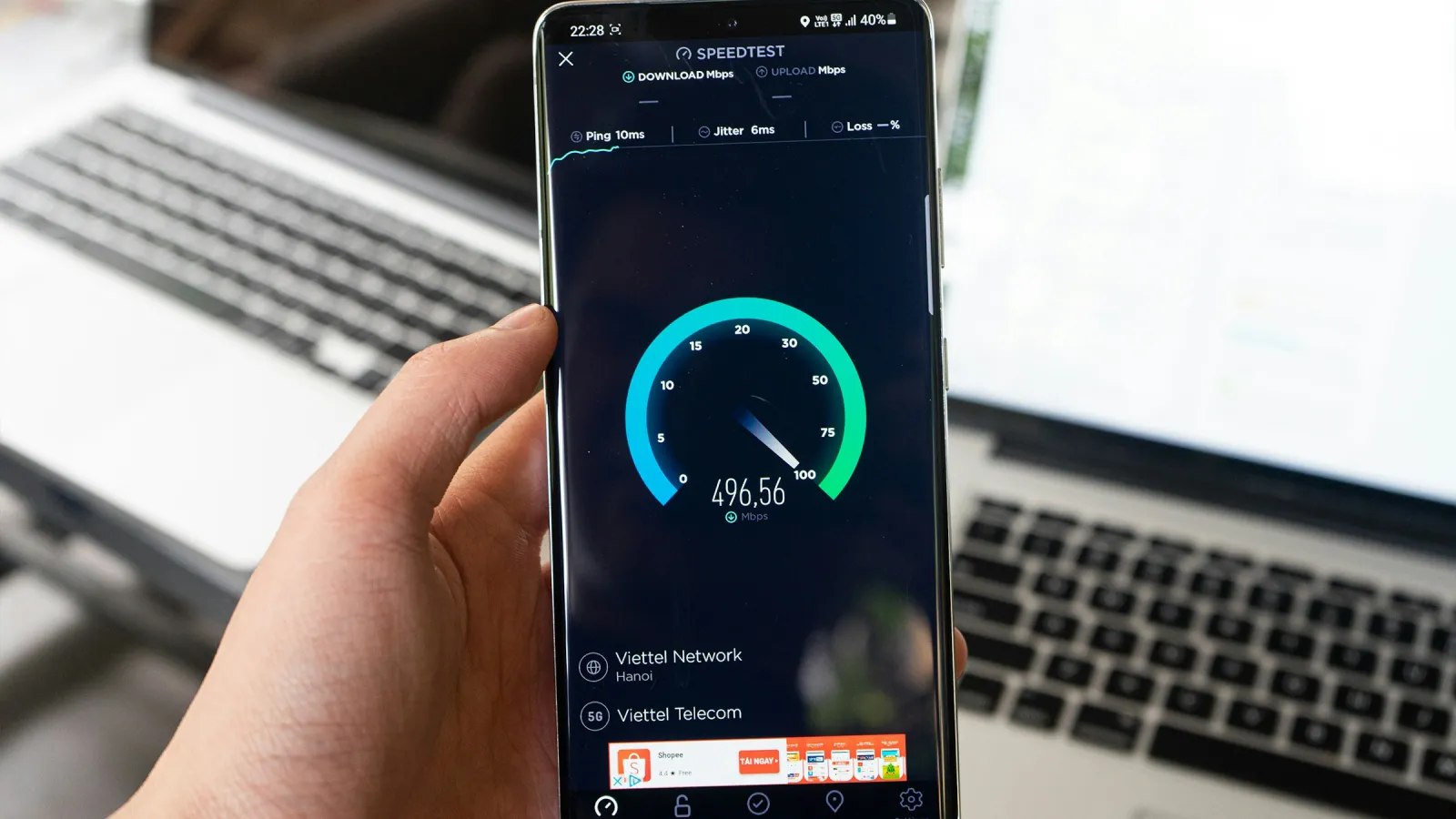 Una mano sujeta un móvil Android que hace un test de velocidad, con un portátil al fondo
